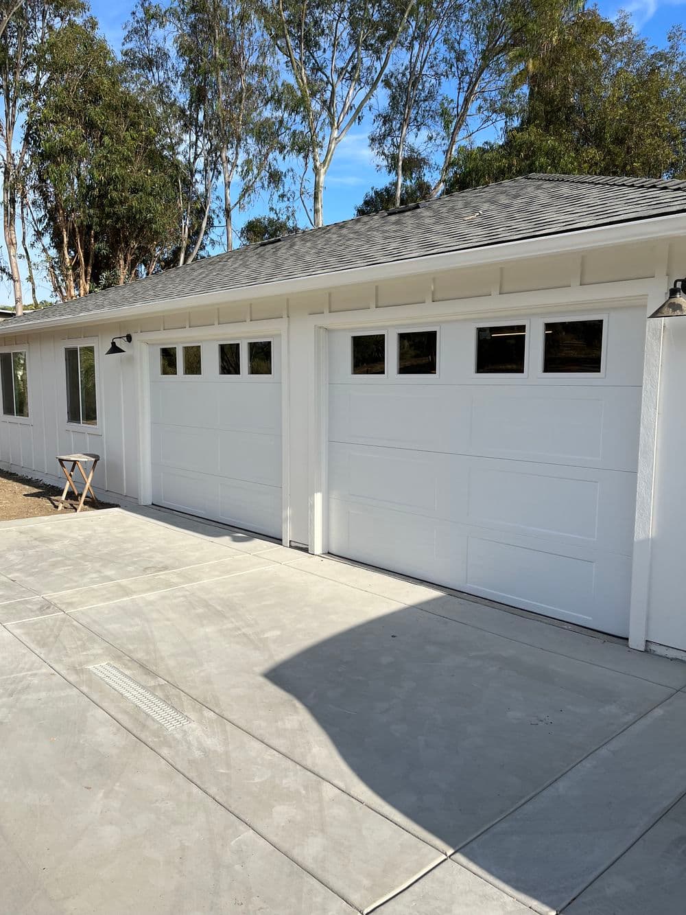Modern white garage doors with windows, surrounded by trees and a newly paved driveway.